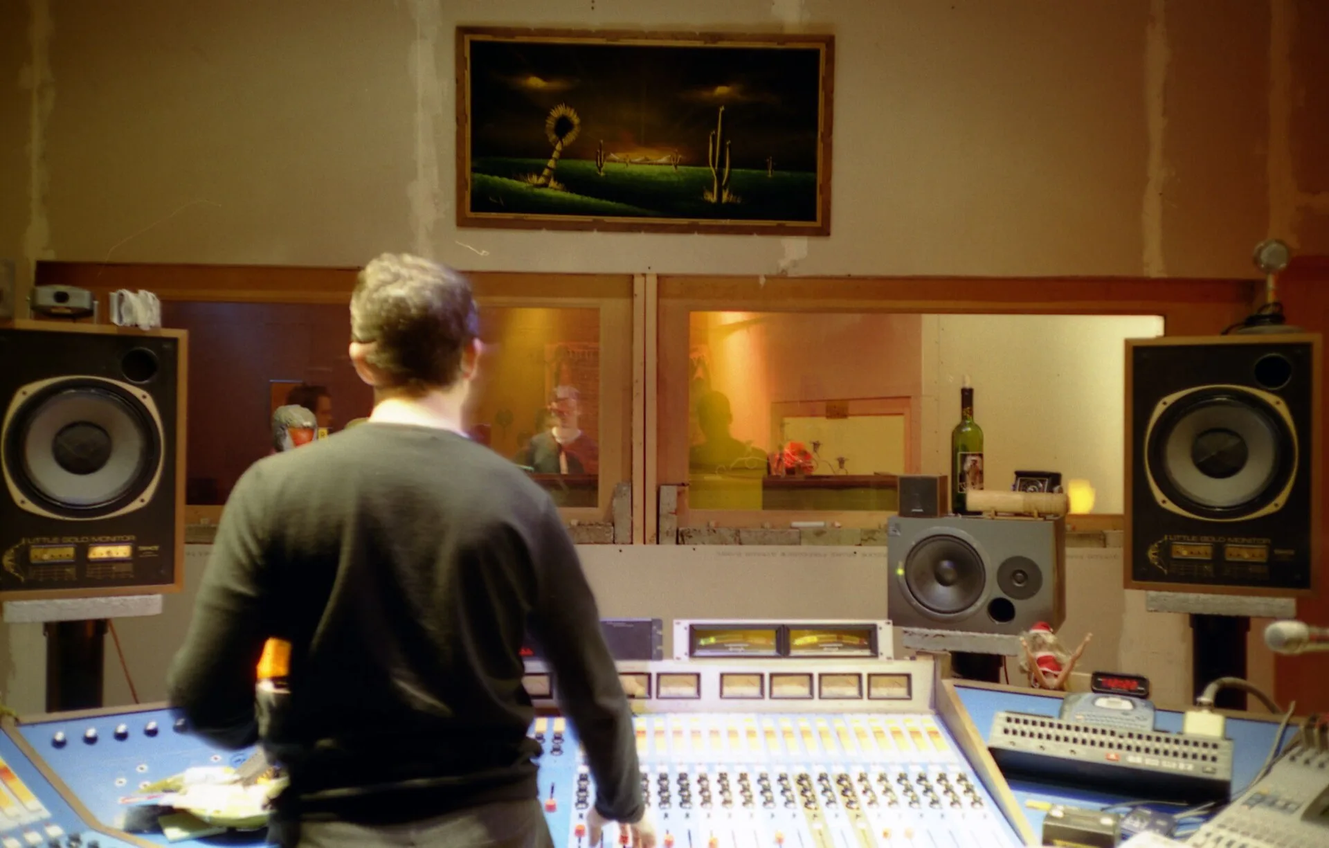 An audio engineer in the control room of a professional sound recording studio. In front of him is the mixing console with a plurality of linear potentiometers for regulating the recording level of the different instruments/voices playing in the live room behind the glass window. In front of the eng. Setting up a desk noise test: what the work involves, explained properly